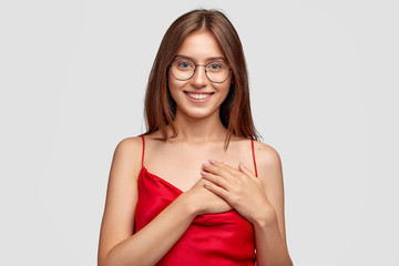 Kind friendly looking young woman with satisfied expression, holds hands on heart, being tactful and thankful for good attitude to her, has broad pleasant smile, stands against white background - obrazy, fototapety, plakaty
