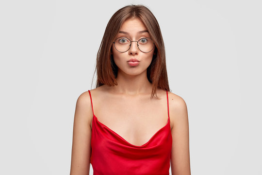 Lovely young lady with friendly expression, keeps lips round, dressed in casual red nightgown, stands against white background. Pleasant girl makes grimace at camera. Facial expressions concept