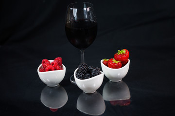 Glass of red wine on a black background with strawberries, blackberries, raspberries