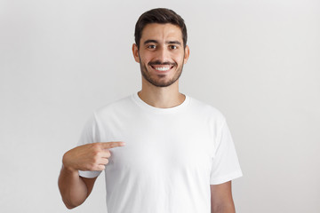 Daylight shot of smiling young man pointing at his blank white t-shirt with index finger, copy...
