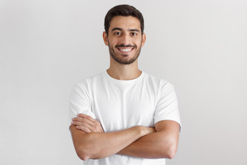 Portrait of smiling handsome man in white t-shirt, standing with crossed arms isolated on gray...
