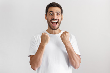Indoor photo of shouting man isolated on gray background, celebrating victory and acting as if he is winner, squeezing fists in deep emotional expression of happiness and luck
