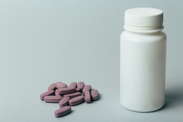 a white box for tablets stands on a gray background, beside lies a bunch of oblong, purple pills.