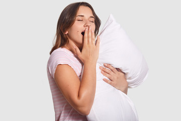 Fatigue woman yawns, keeps hand on opened mouth, wants to sleep after sleepless night, holds pillow, dressed in nightwear, poses against white background. People, rest and lifestyle concept.