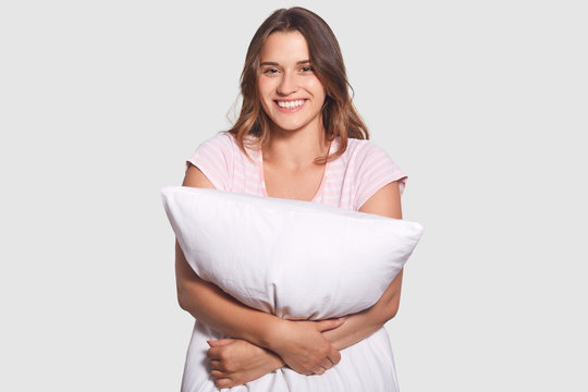 Cheerful healthy young woman with toothy smile, being in good mood after awakening and having pleasant dreams, holds pillow, poses in bedroom, has appealing look, isolated over white background
