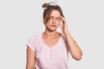 Fatigue woman cant get asleep, has terrible headache, keeps hand on temple, dressed in casual pyjamas, closes eyes from pain, isolated over white background. Weariness and tiredness concept.