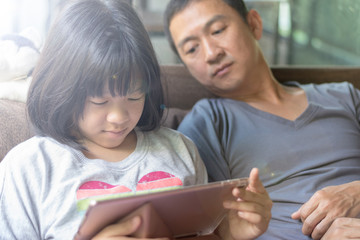 Fototapeta na wymiar Father and daughter activity technology together concept. Asia kid girl reading e-book or playing game on computer tablet while dad watching as parental guidance for PG13.