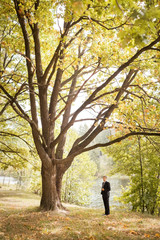 The groom in a black suit is standing by a large old oak tree in the park on the background of a pond