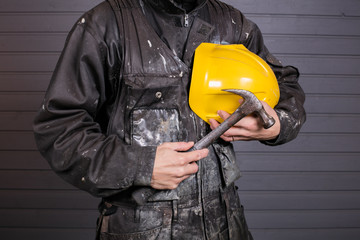 The workman holds in his hand the yellow protective helmet and hammer. The Worker overalls are dirty.
