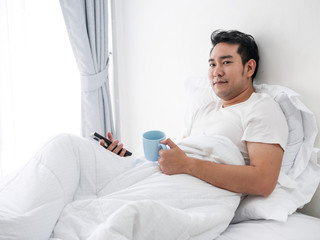 Obraz premium Smiling Asian man using smartphone and drinking coffee on bed, man lifestyle on weekend concept.