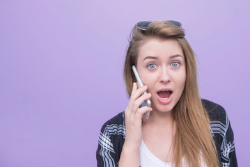 Emotional girl talking on the phone and looking at the camera on a purple background. Portrait of a surprised young woman talking on a smartphone and isolated purple background.