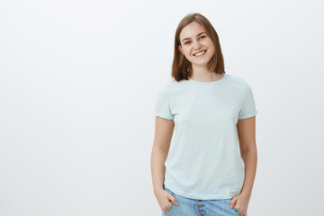 Friendly cute white girl with short haircut and lovely flapped ear smiling joyfully holding hands in pockets and gazing at camera with joyful entertained look posing over white background
