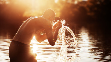 a young man is standing in a river and washing his face against the sunset.