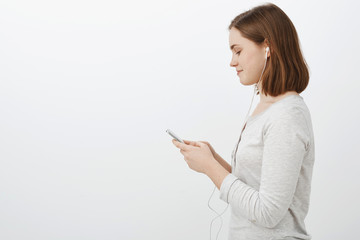 Naklejka premium Profile shot of cute sociable young caucasain female brunette wearing earphones standing against grey wall in casual white blouse holding smartphone smiling while texting friend on her way to party