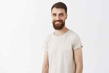 Fototapeta premium Happy creative and young bearded male coworker with stylish hairstyle in white casual t-shirt smiling joyfully standing over gray background talking about new company concepts creating new ideas