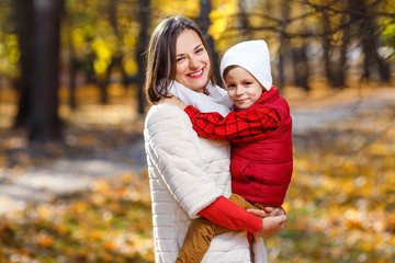 Cute,happy, white boy in red shirt smiling and hugging with his mom among yellow leaves. Little child having fun with mother in autumn park. Concept of friendship between son and parents, happy family