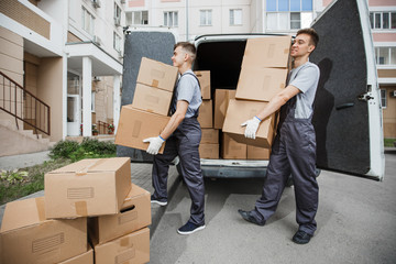 Two young handsome smiling workers wearing uniforms are unloading the van full of boxes. The block of flats is in the background. House move, mover service.