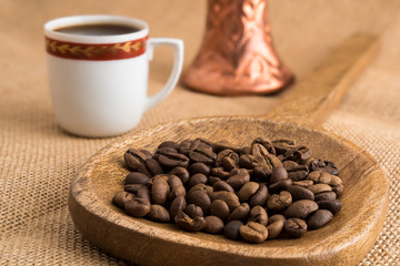 Roasted coffee beans in wooden ladle and traditional bronze coffee pot and cup on sackcloth