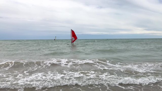 Windsurfers fall on sea waves. Windsurfer in buoyancy aid on windsurf with red sail on waves falls to the aquamarine water, windy dangerous day, cloudy low sky