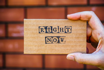 Woman hand holding cardboard card with words Right Now against brick wall background.