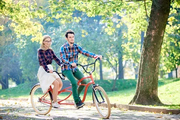 Tuinposter Young active smiling traveler couple, handsome bearded man and attractive blond woman riding together tandem double bike along path in lit by bright sun beautiful park under tall trees. © anatoliy_gleb