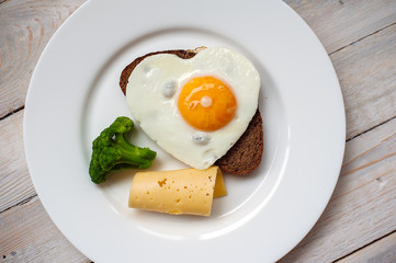 The fried egg in the form of heart with vegetables and bread on a white plate