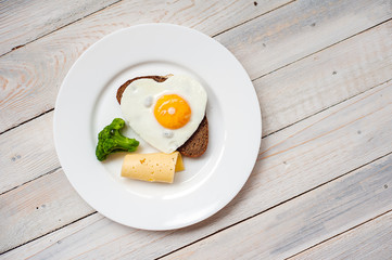 The fried egg in the form of heart with vegetables and bread on a white plate