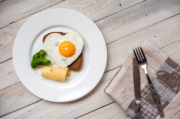 The fried egg in the form of heart with vegetables and bread on a white plate