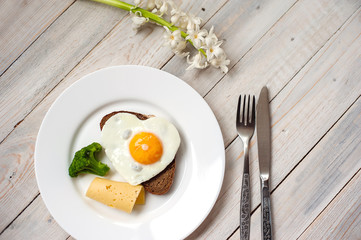 The fried egg in the form of heart with vegetables and bread on a white plate