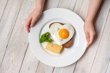 The fried egg in the form of heart with vegetables and bread on a white plate