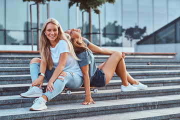 Portrait of a two young hipster girls sitting together on skateboard at steps on a background of...