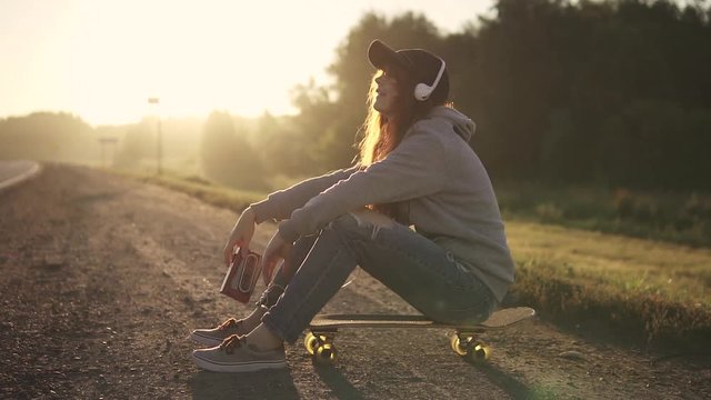 portrait of a girl sitting on a sketchboard on the roadside and listening to music on headphones. teenager and youth culture