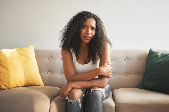Indoor shot of beautiful fashionable young mixed race woman with Afro hairstyle sitting on couch at home, frowning, having worried sad look, suffering from stomach cramps or feeling lonely and upset