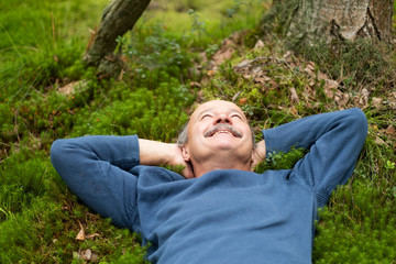 Mature man in blue sweater lay on grass in the a forest listening to birds songs.