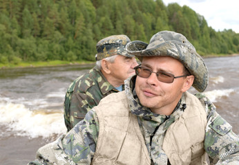 Two fisherman are wearing camouflage riding a motor boat on a river. The happy man, 30 years old, and the elderly man, 80 years old, are traveling on a water vehicle. The grandfather and his grandson.