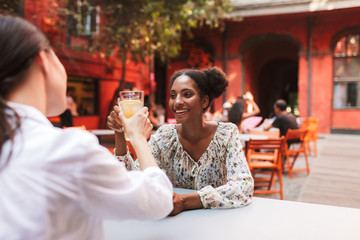 Pretty smiling african girl in dress drinking cocktails with friend while joyfully spending time together in cozy courtyard of cafe