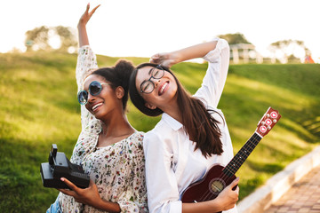 Cheerful girls with little guitar and polaroid camera in hands joyfully spending time in park