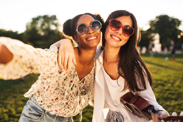 Two beautiful girls in sunglasses friendly hugging each other while happily taking photos together in city park