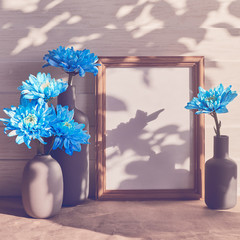 Blue chrysanthemums are found in a variety of mini-vases. Nearby there is an empty wooden frame. White wooden background.