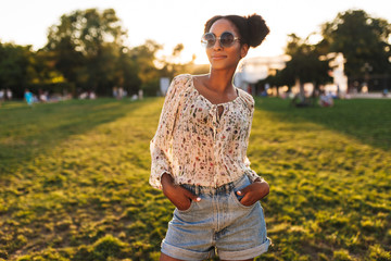 Fototapeta premium Young beautiful african woman in sunglasses dreamily looking in camera while spending time in city park