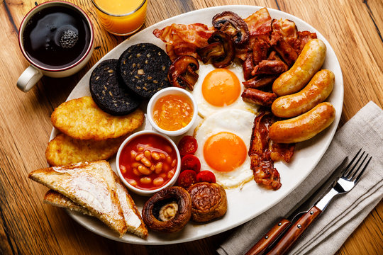 Full fry up English breakfast with fried eggs, sausages, bacon, black pudding, beans, toasts and tea on wooden background