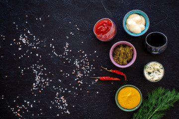 Different dip sauce in bowls near chili pepper and greenery. Ketchup, mayonnaise, mustard, soy sauce, barbecue sauce, pesto, mustard, sour on black background top view copy space