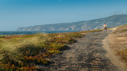 Naklejka premium Mature man (55-60) cycles along a bumpy asphalt path overlooking Praia do Guincho and Roca do Cabo near Cascais, Portugal during a windy summer day