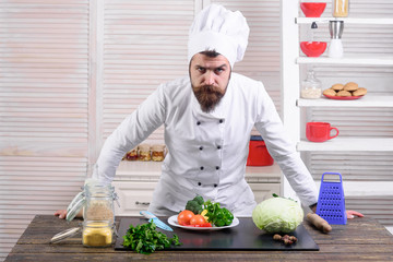 Bearded serious chef stands in kitchen near table. Cook in white uniform stands on restaurant kitchen. Master chef. Cooking, culinary, cuisine. Vegetarian, health, meal, diet. Food preparation concept