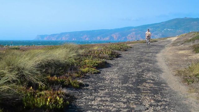 Mature man (55-60) cycles along a bumpy asphalt path overlooking Praia do Guincho and Roca do Cabo near Cascais, Portugal during a windy summer day
