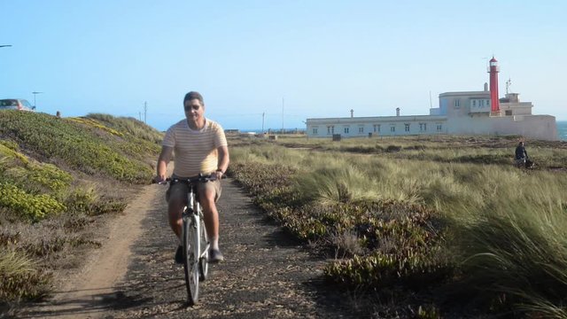 Mature man (55-60) cycles along a bumpy asphalt path overlooking the lighthouse in Cape Raso, Cascais - Portugal
