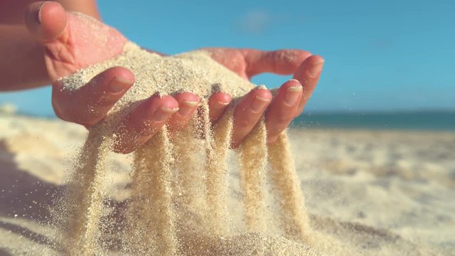 SLOW MOTION, CLOSE UP: Unrecognizable female tourist holds up her hands and scatters the hot sand across the tranquil shore. Awesome shot of white particles of sand slipping through woman's fingers.