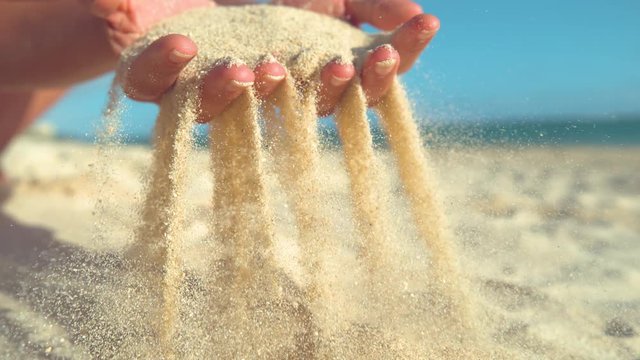 SLOW MOTION, CLOSE UP: Unknown young woman scattering white sand between her gentle fingers. Playful female tourist scooped up a handful of sand and lets it get blown away in the cool ocean breeze.