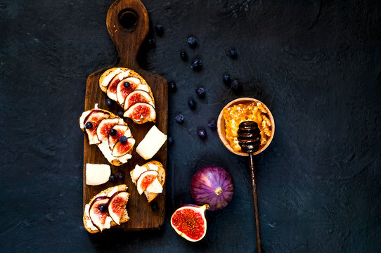 Sandwiches with fresh figs, goat cheese, honey and blueberries on wooden board.Vegetarian food.Creative layout of the whole and sliced figs on dark blue background. Flat lay. Copy space, closeup.