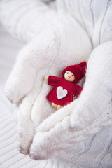 Hands in white knitted mittens holding little red Christmas doll with heart.